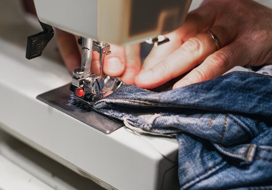 Detailansicht einer Nähmaschine mit Händen, welche eine Jeanshose reparieren