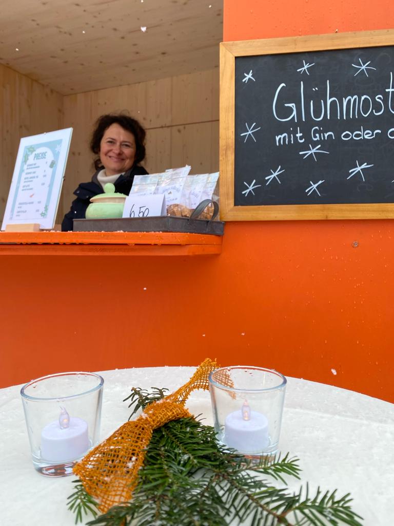 Eine Frau verkauft Glühmost in einem orangen Holzhäuschen. Im Vordergrund liegen Tannzweige und Kerzen 